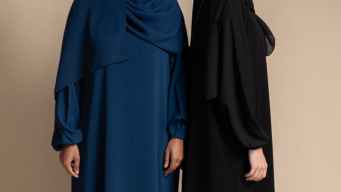 Two women wearing long, flowing abayas in blue and black on a beige background