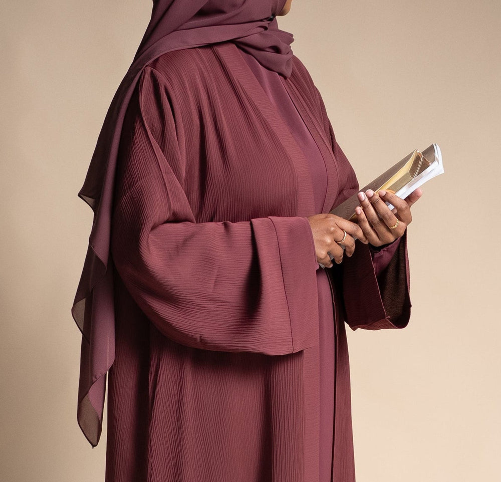 Person wearing a long burgundy dress and hijab holding a book against a beige background