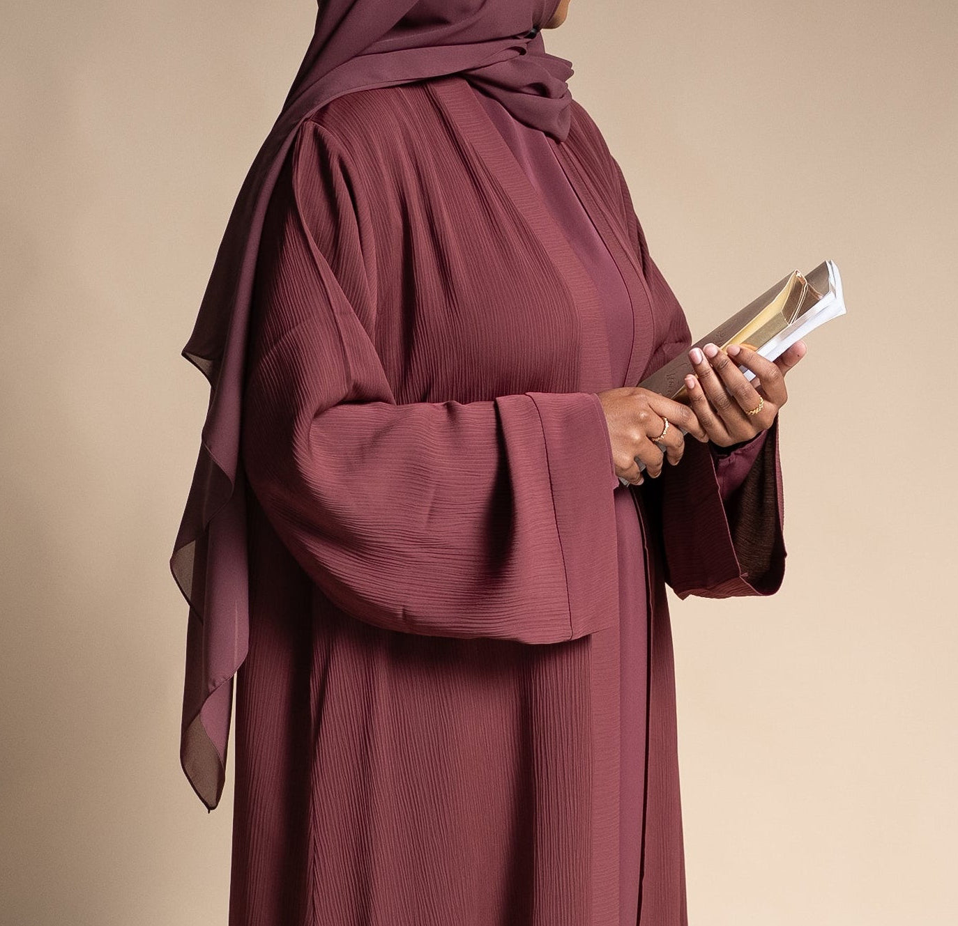 Person wearing a long burgundy dress and hijab holding a book against a beige background