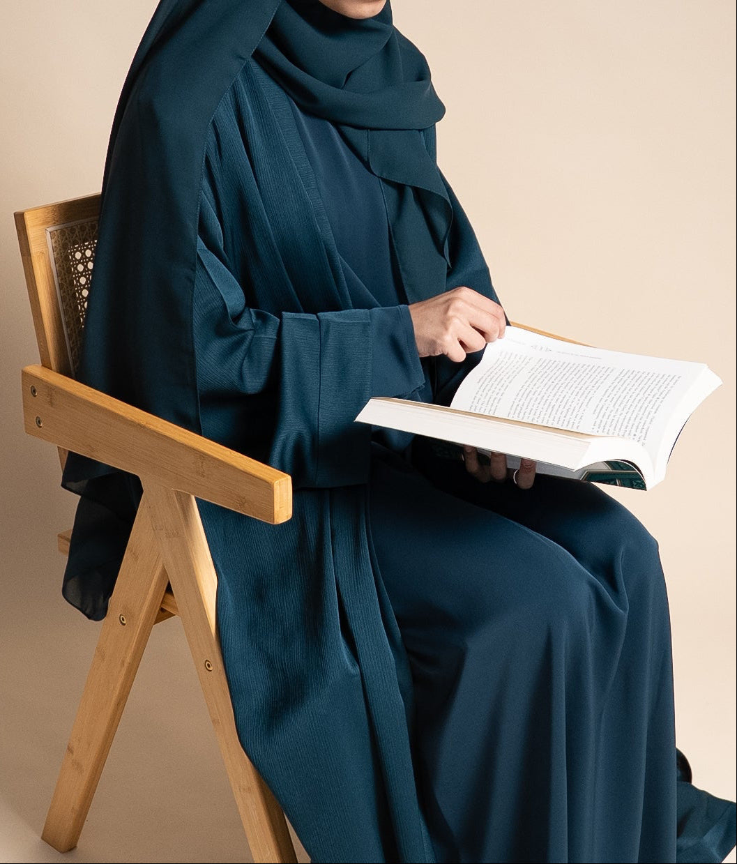 Person in a teal abaya sitting on a wooden chair holding an open book against a beige background