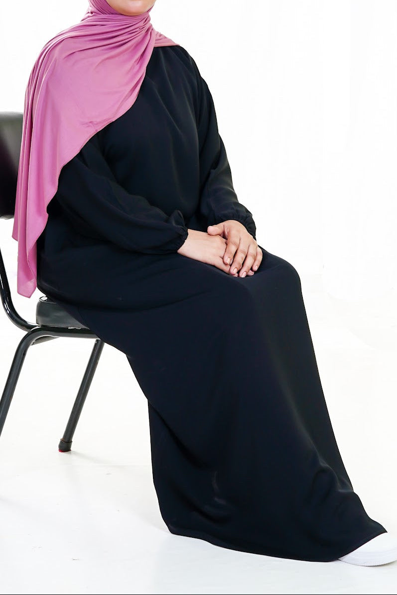 Person wearing a black abaya with a pink hijab sitting on a chair.