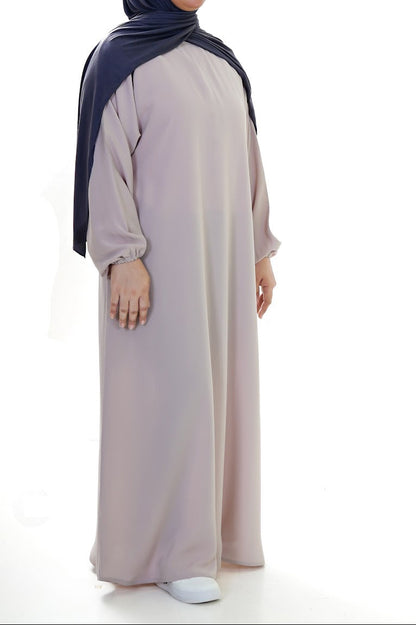 Woman wearing a long, stone beige abaya with a dark hijab on a white background