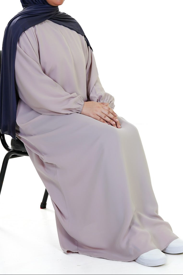 Person wearing a long, stone beige abaya sitting on a chair against a white background