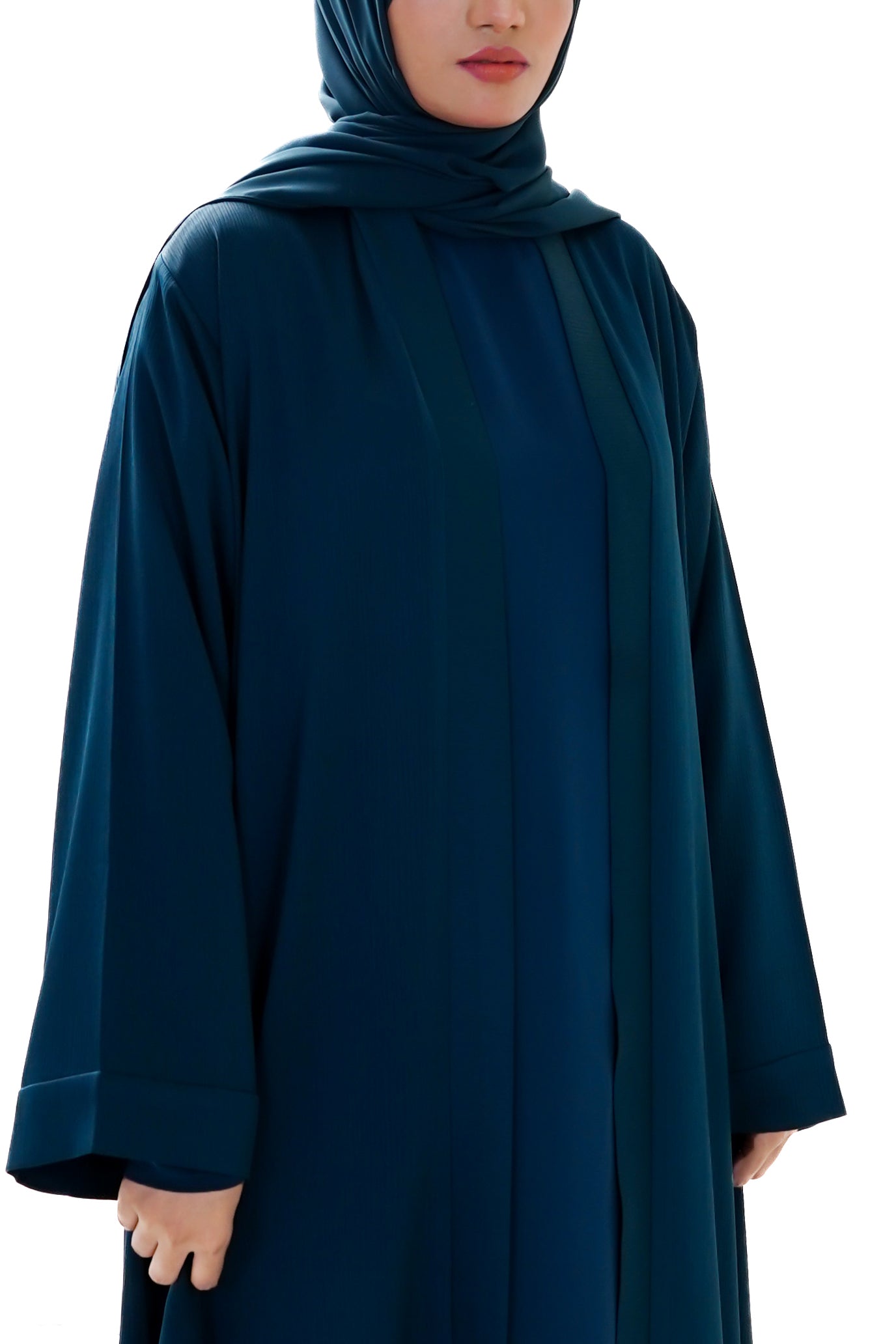 Woman wearing a long deep teal abaya with a matching hijab on a white background