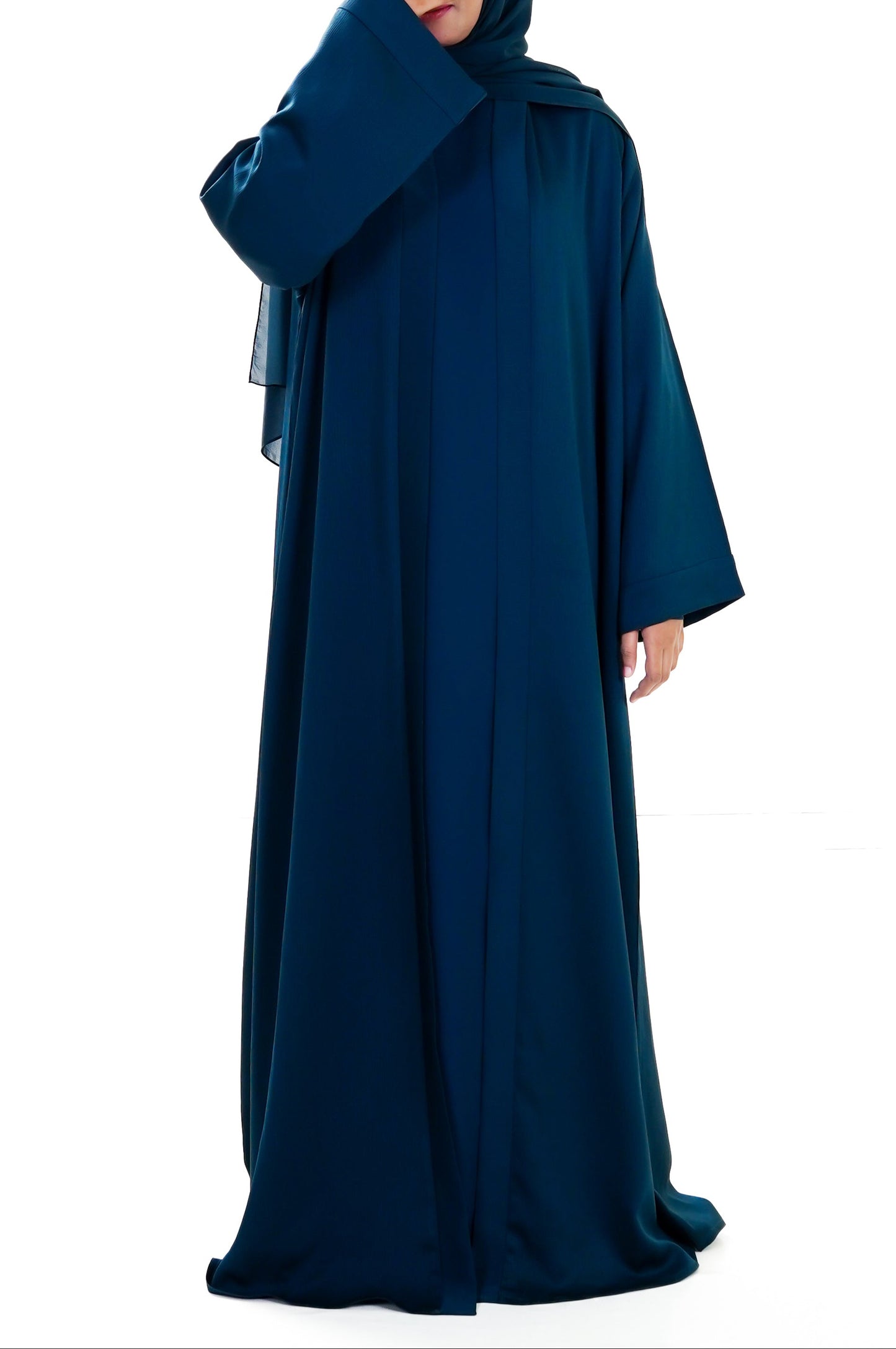 Woman wearing a long deep teal abaya with a matching hijab on a white background