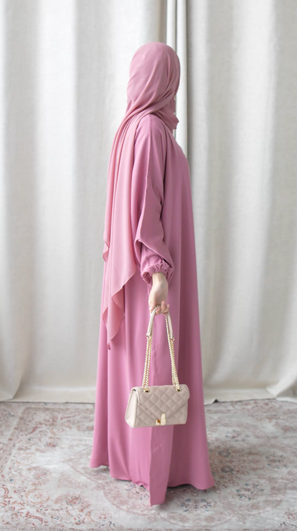 Person wearing a pink hijab and holding a white handbag against a white curtain background
