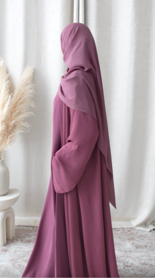 Person wearing a long, dark purple hijab standing in a room with white curtains and decorative plants.